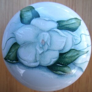 May include: A white ceramic cabinet knob with a hand-painted floral design. The design features a large white flower with green leaves and a light blue background. The knob is round and has a glossy finish.