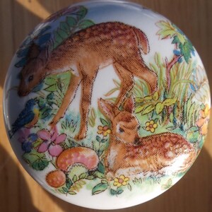 May include: A round, white ceramic decorative piece featuring a detailed illustration of two deer in a meadow. The deer are brown with white spots, surrounded by colorful flowers, green foliage, and a blue bird. The piece has a glossy finish.