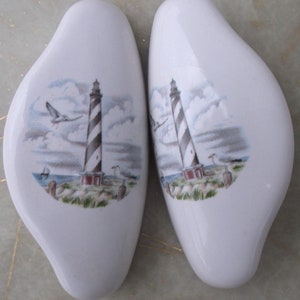 May include: Pair of white ceramic lighthouse salt and pepper shakers. Each shaker features a black and white striped lighthouse with a red roof, a blue sky with white clouds, and a seagull flying overhead.