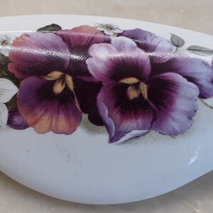 May include: White, oval-shaped decorative object with a floral design. The design features purple pansies with yellow centers, along with white and gray flowers. The object has a glossy finish.