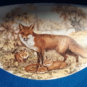 May include: A white, curved ceramic piece featuring a detailed illustration of a family of foxes in a woodland setting. The artwork depicts a red fox standing with three cubs, surrounded by foliage and trees.