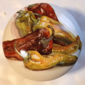 May include: A round, white decorative knob featuring a colorful illustration of various chili peppers. The peppers are depicted in shades of red, yellow, and green, with detailed shading and highlights. The knob is likely made of ceramic or porcelain.
