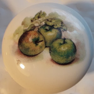 May include: A white ceramic knob with a painted image of three green apples with leaves.