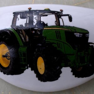 May include: White, oval-shaped decorative stone featuring a green and black tractor with yellow wheels. The tractor design is detailed, showcasing the vehicle's features. A unique decorative item for farm enthusiasts.
