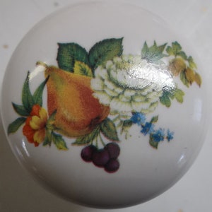 May include: A white ceramic knob with a floral design featuring a pear, a white rose, and cherries. The floral design is in shades of yellow, orange, green, white, and blue.