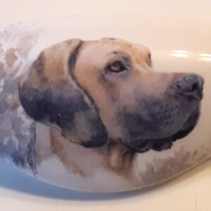 May include: A ceramic, oval-shaped decorative piece featuring a painted portrait of a dog. The dog has tan and black fur, with a detailed depiction of its face and ears. The background is a soft, watercolor-like wash.