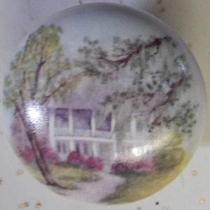 May include: A hand-painted ceramic ornament featuring a white house with a porch and trees in the background. The house is painted in shades of purple and white, and the trees are painted in shades of green and brown.