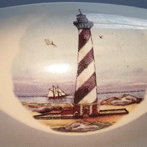 May include: A ceramic decorative piece with a lighthouse scene. The lighthouse is painted with alternating red and white stripes, set against a coastal landscape with a sailboat and birds. The piece has a light blue border.
