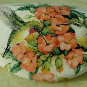 May include: A white, oblong decorative object featuring a detailed illustration of two hummingbirds hovering near a cluster of orange flowers with green leaves. The birds have green and yellow plumage.