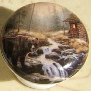 May include: A ceramic round trinket box with a woodland scene featuring a brown bear walking by a stream in a forest. A small cabin with a lit window is in the background.