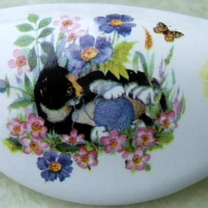 May include: White ceramic decorative piece with a painted design of a cat playing with yarn amidst flowers and butterflies. The piece has a curved, elongated shape, with a cat and fishbowl design on each end.