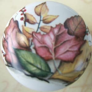 May include: A white ceramic cabinet knob featuring a painted design of autumn leaves. The leaves are in shades of red, green, and yellow, with small red berries. The knob is round and has a glossy finish.