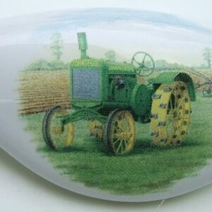 May include: A white ceramic decorative piece featuring a green and yellow tractor in a field. The tractor has large yellow wheels and is set against a backdrop of a field and sky. The piece is likely a decorative item.