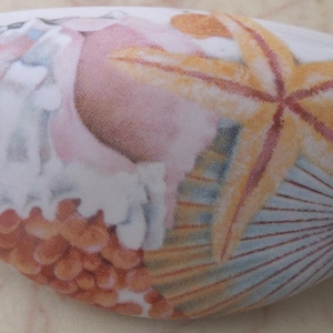May include: A white ceramic oval-shaped dish with a painted design of a seashell, starfish, and a fan-shaped shell. The seashell is white with pink and orange accents. The starfish is orange and yellow. The fan-shaped shell is blue and white.