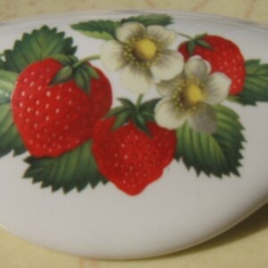 May include: White ceramic drawer pull with a strawberry design. The design features red strawberries, green leaves, and white flowers with yellow centers. The pull has a curved, ergonomic shape.