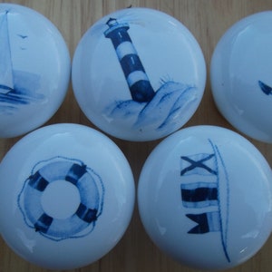 May include: Five white ceramic cabinet knobs with nautical designs in blue. The designs include a sailboat, a lighthouse, an anchor, a life preserver, and nautical flags. The knobs are round and have a glossy finish.