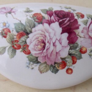 May include: White ceramic drawer pull with a floral design. The design features pink and burgundy roses, red strawberries, and green leaves. The drawer pull is oval-shaped and has a smooth, glossy finish.