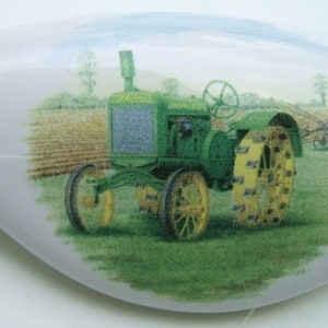 May include: A green and yellow tractor with a plow in a field. The tractor has a large front tire and a smaller rear tire. The tractor is in a field with green grass and brown dirt.