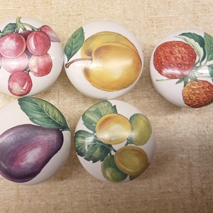 May include: Five white ceramic cabinet knobs with hand-painted fruit designs. The knobs feature illustrations of plums, an apricot, a strawberry, a plum, and apricots. Each knob has a different fruit design with green leaves.