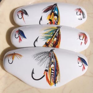 May include: Three white ceramic fly fishing lures with colorful fly patterns. Each lure has a hook and a different fly pattern.
