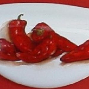 May include: A white, oblong ceramic dish with a slight curve, holding several bright red chili peppers. The peppers have green stems and are arranged in a cluster. The dish sits on a red surface.