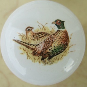 May include: A white ceramic knob with a brown and green pheasant design. The pheasant is depicted in a natural setting with brown and green foliage.