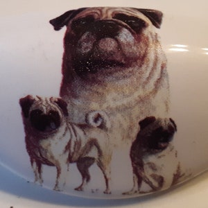 May include: A white ceramic heart-shaped dish with a printed image of three pugs. The pugs are brown and fawn with black noses and eyes. The largest pug is standing and the two smaller pugs are sitting.