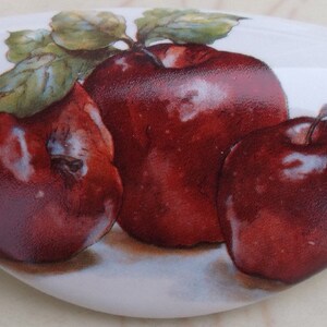 May include: A white, oval-shaped decorative ceramic piece featuring a painting of three ripe red apples with green leaves. The apples have highlights and shadows, giving them a realistic appearance.