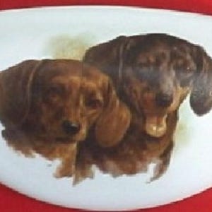 May include: White, elongated ceramic dish featuring a detailed painting of two dachshunds. The dogs are depicted in shades of brown and black, with expressive faces. The dish has a smooth, glossy finish and is set against a red background.