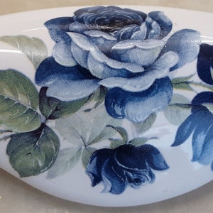 May include: White, oval-shaped decorative object with a floral design. The design features blue roses and green leaves. The object has a glossy finish and appears to be made of ceramic or porcelain.