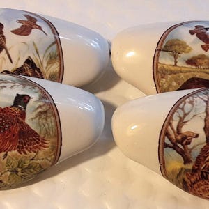 May include: Four white ceramic drawer pulls with oval shapes. Each pull features a different detailed illustration of birds in a natural landscape, including pheasants and other game birds. The artwork is framed by a brown border.