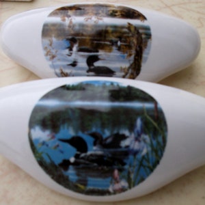 May include: Two white ceramic drawer pulls with a round, colorful image of a lake scene with birds and trees. The top pull features a loon swimming in the lake. The bottom pull features a loon with its head above the water.