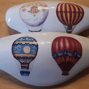 May include: Two white oval cabinet knobs with colorful hot air balloon designs. One knob features a yellow and purple balloon, while the other displays a red and yellow striped balloon. The knobs are decorative and suitable for furniture.