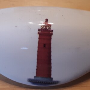 May include: White ceramic dish with a red and white lighthouse design. The dish has a rounded shape and a smooth surface.