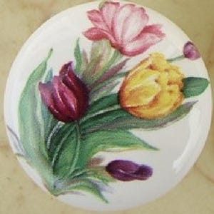 May include: A round, white ceramic decorative knob featuring a hand-painted floral design. The design includes pink, yellow, and purple tulips with green leaves. The knob is likely for a cabinet or drawer.