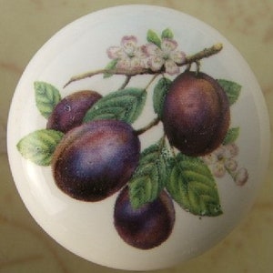 May include: A white ceramic knob with a floral design featuring purple plums and green leaves. The knob has a white background with a floral design in the center.