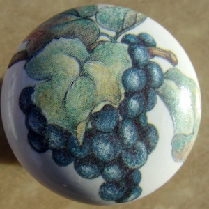 May include: A white ceramic knob with a grapevine design. The grapes are a dark blue color and the leaves are green and yellow.