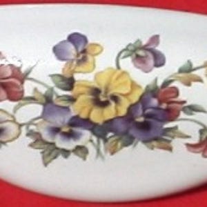 May include: A white, oblong ceramic dish featuring a floral design of pansies in shades of yellow, purple, and burgundy. The pansies are arranged with green leaves against a white background. The dish has a smooth, glossy finish.