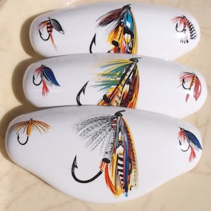 May include: Three white ceramic oval-shaped ornaments with colorful fly fishing lures painted on them. Each ornament features a different lure design with a variety of colors, including red, yellow, blue, green, and black.