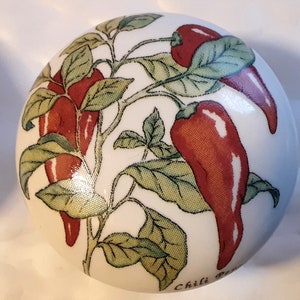 May include: A white ceramic knob featuring a design of red chili peppers and green leaves on a vine. The knob has a glossy finish and the words "Chili Peppers" are printed on the bottom. This decorative knob is suitable for kitchen or dining room decor.