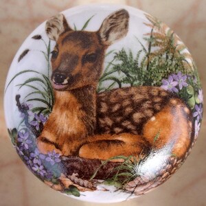 May include: A round, white ceramic knob featuring a detailed illustration of a fawn resting in a field of purple flowers and green foliage. The fawn has brown fur with white spots and is lying down. The knob is likely for a cabinet or drawer.
