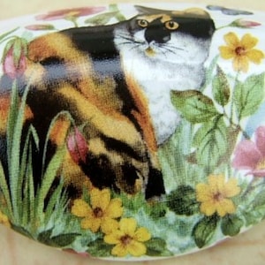 May include: White oval ceramic cabinet knob featuring a calico cat amidst colorful flowers and greenery. The cat has black, orange, and white fur. Yellow and pink flowers are scattered around the cat, with green leaves and grass.