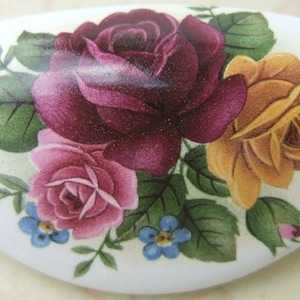 May include: White, oval-shaped decorative ceramic piece with a floral design. The design features a cluster of roses in shades of burgundy, pink, and yellow, along with green leaves and small blue flowers. The piece has a glossy finish.