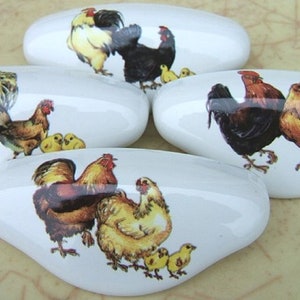 May include: Five white, oval-shaped ceramic cabinet knobs. Each knob features a different illustration of a rooster, hen, and chicks in various colors. The knobs are arranged on a light brown surface.