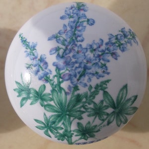 May include: A white, round ceramic cabinet knob featuring a floral design. The design includes blue delphinium flowers and green leaves. The knob is a decorative element for furniture and home decor.