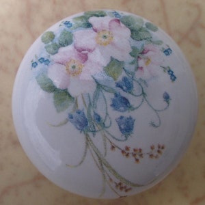 May include: A white ceramic knob with a floral design. The design features pink and white flowers with green leaves and blue bell-shaped flowers.