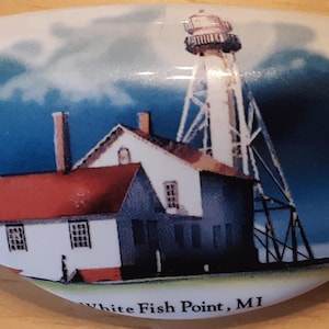 May include: White ceramic drawer pull featuring a detailed illustration of the White Fish Point Lighthouse in Michigan. The image shows the lighthouse, a house with a red roof, and the text "White Fish Point, MI".