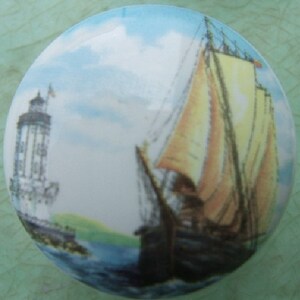 May include: A ceramic knob with a painted image of a sailboat with white sails and a lighthouse in the background.