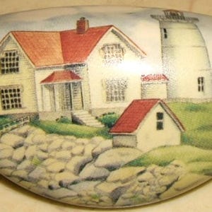 May include: A white, oval-shaped decorative item featuring a painted coastal scene. The artwork depicts a lighthouse, a house, and a small building with red roofs, set against a rocky shoreline and green grass.