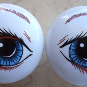 May include: Two white ceramic knobs with blue eyes and brown eyelashes. The eyes have black pupils and a blue iris.